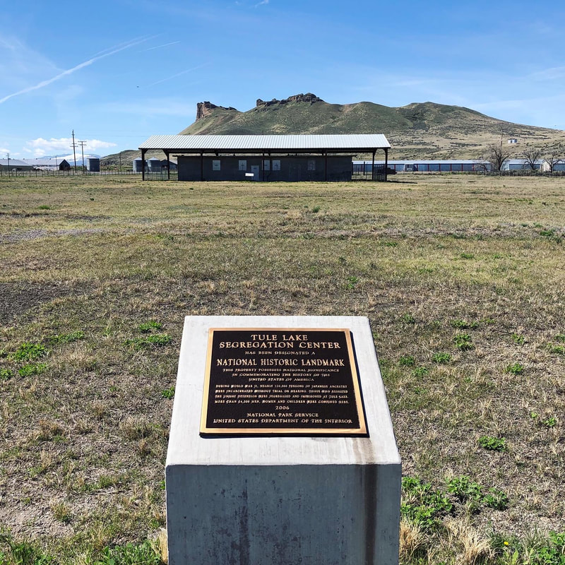 A memorial plaque on a dry grassy plain with a squat concrete building beneath an awning and a rugged mountain in the background